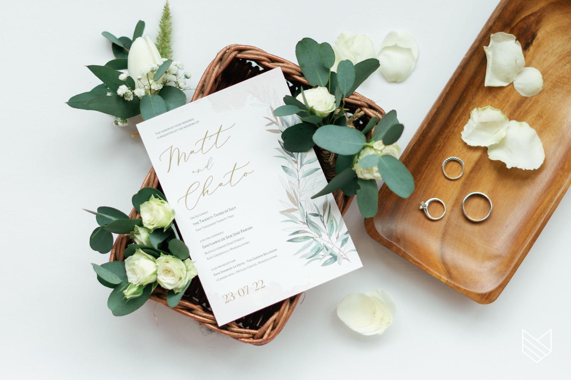 A wedding invitation suite in gold script, styled with eucalyptus, white roses and two rings on a wooden tray.